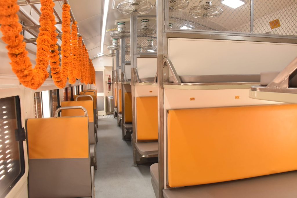Interior view of the General Second Class coach in the Banaras-Pune Amrit Bharat Express featuring orange and grey cushioned seats, overhead luggage racks decorated with marigold garlands, and aero-dynamic ceiling fans.