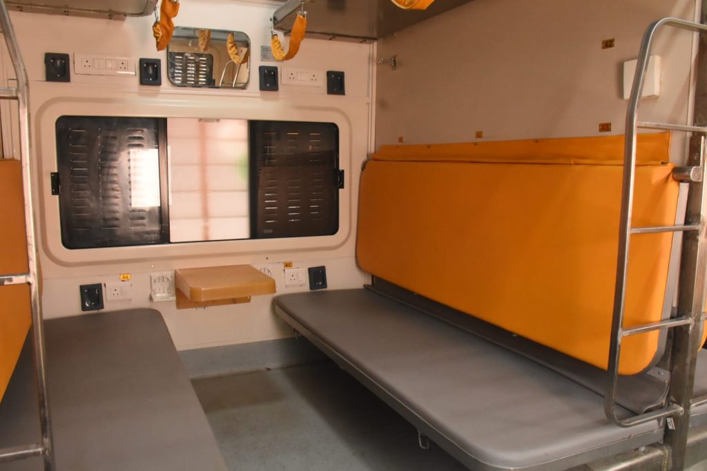 Interior of a Sleeper Class compartment in the Banaras-Pune Amrit Bharat Express showing wide berths with orange padding, integrated mobile charging points, foldable snack tables, and improved ladder design for upper berths.