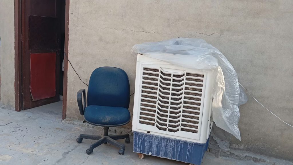 A white and blue desert air cooler stands next to a blue office chair against a plain wall. The cooler is partially covered with a clear plastic sheet, and its water tank shows visible white mineral buildup and water stains.