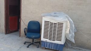 A white and blue desert air cooler stands next to a blue office chair against a plain wall. The cooler is partially covered with a clear plastic sheet, and its water tank shows visible white mineral buildup and water stains.