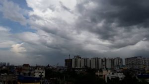 A wide-angle shot of the Pune skyline under a heavy, overcast sky filled with dark, dramatic storm clouds, signaling an approaching thunderstorm and light rain.