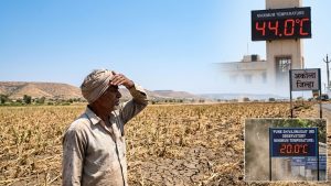 A wide, realistic documentary photograph capturing the severe heatwave in Maharashtra. In the foreground, deeply cracked, parched agricultural land in Vidarbha dominates the scene. A man, shielding his face from the intense sun, looks over the desolate landscape. In the background, rolling sun-bleached hills stretch to a shimmering horizon under a clear, bright blue sky. A distant digital temperature display on a tower in Akola reads '44.0°C' in red LEDs, with the Marathi sign for Akola District (अकोला जिल्हा) nearby. Heat haze distorts the horizon, creating a palpable sense of extreme heat and aridity.