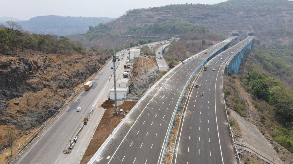 An aerial shot showing the old winding ghat road on the left contrasted with the new, straight multi-lane "Missing Link" bypass and its towering blue-pillared viaduct on the right.