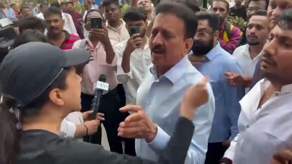 An intense close-up shot from a viral video showing a woman in a black baseball cap and t-shirt confronting Maharashtra Minister Girish Mahajan on a crowded Mumbai street. The woman is gesturing emphatically with her finger while speaking to the minister, who wears a light blue shirt and appears to be responding amidst a crowd of onlookers, supporters, and media personnel holding microphones and smartphones.