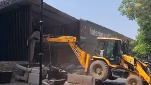 A yellow JCB excavator demolishes a large illegal commercial structure with a grey facade and "Interior and Design" signage during a PCMC anti-encroachment drive in Pimpri-Chinchwad. Debris and furniture are visible on the ground in the foreground under a clear sky.