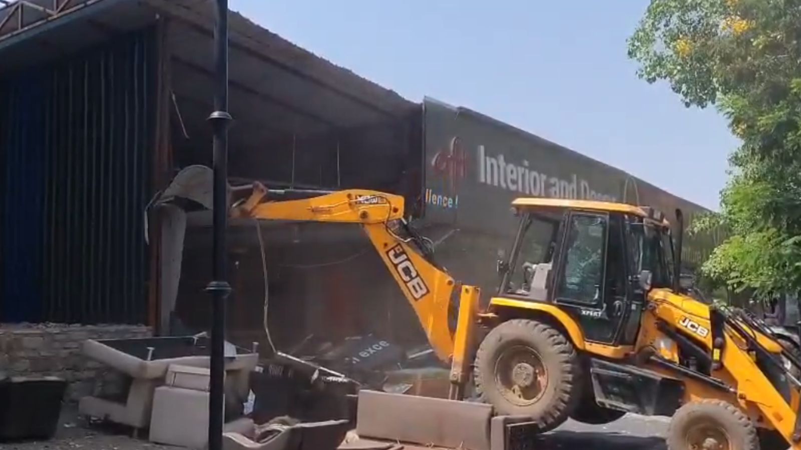A yellow JCB excavator demolishes a large illegal commercial structure with a grey facade and "Interior and Design" signage during a PCMC anti-encroachment drive in Pimpri-Chinchwad. Debris and furniture are visible on the ground in the foreground under a clear sky.