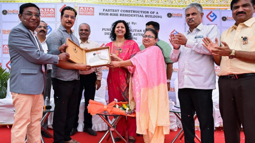 A group of officials standing on a stage, holding an official certificate and a plaque to celebrate the record-breaking construction of a high-rise residential building by the PMRDA. A large banner in the background displays the logos for the Asia Book of Records and the India Book of Records.