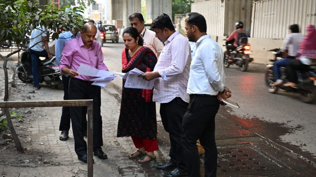 PMRDA Additional Commissioner K. Manjulakshmi and a team of senior officials and engineers conduct a site inspection for Pune Metro Line 3. The group is standing on a sidewalk near University Circle, reviewing technical documents and blueprints on-site to resolve construction bottlenecks. Traffic and metro pillars are visible in the background.
