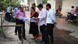 PMRDA Additional Commissioner K. Manjulakshmi and a team of senior officials and engineers conduct a site inspection for Pune Metro Line 3. The group is standing on a sidewalk near University Circle, reviewing technical documents and blueprints on-site to resolve construction bottlenecks. Traffic and metro pillars are visible in the background.