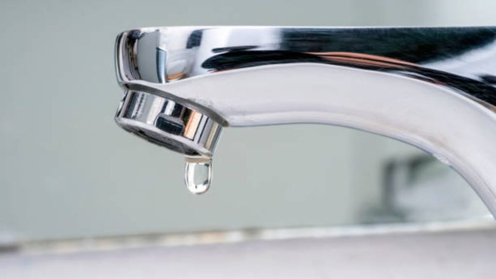 A close-up of a chrome faucet with a single water droplet hanging from the spout, symbolizing a water shortage or supply disruption.