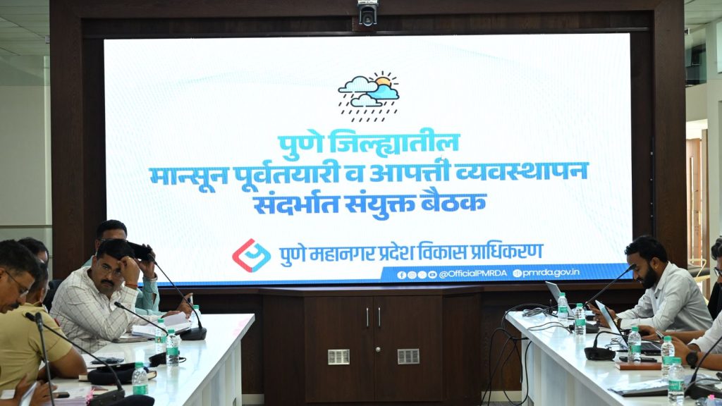 A meeting room with PMRDA officials seated at tables on either side. A large digital screen in the background displays the text "Joint Meeting on Pre-monsoon Preparation and Disaster Management in Pune District" in Marathi beneath a cloud and rain icon.