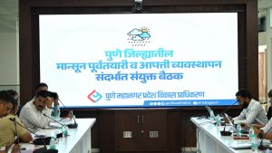 A meeting room with PMRDA officials seated at tables on either side. A large digital screen in the background displays the text "Joint Meeting on Pre-monsoon Preparation and Disaster Management in Pune District" in Marathi beneath a cloud and rain icon.
