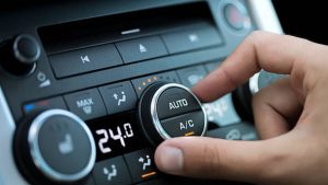 Close-up of a hand adjusting a car's climate control dial, with the digital display clearly showing the temperature set to 24.0°C. The focus is on the "AUTO" and "A/C" buttons on the central knob, highlighting the recommended setting for balancing cabin cooling and fuel efficiency during extreme heat.