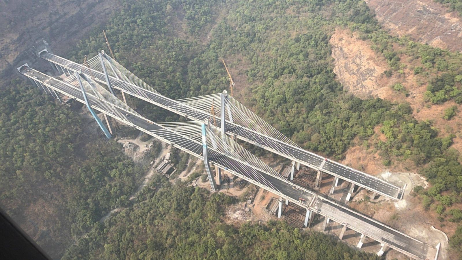 An aerial high-angle view of the massive under-construction cable-stayed bridge of the Mumbai-Pune Expressway "Missing Link" project, spanning deep through the lush green forests of Tiger Valley near Lonavala, featuring twin multi-lane roadways emerging from tunnels into the bridge structure.