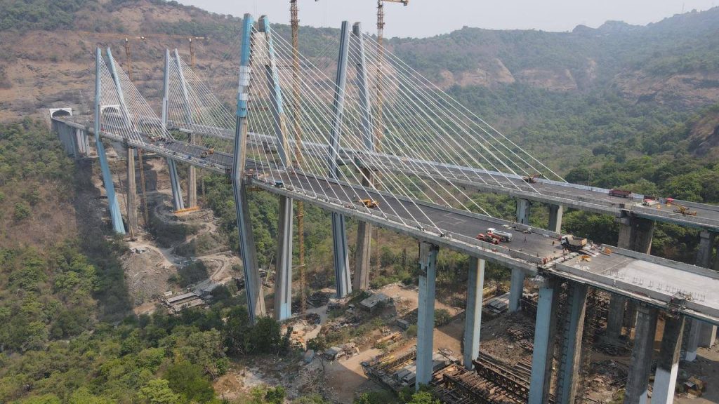 A side-profile view of the towering cable-stayed bridge in Tiger Valley, featuring high blue and white pylons and dense networks of stay-cables supporting the road deck high above the valley floor.