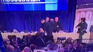 High-angle shot of a security incident at the 2026 White House Correspondents' Dinner. Inside a dimly lit, crowded ballroom at the Washington Hilton, several Secret Service agents in dark suits are gathered around the head table, where President Donald Trump is being quickly escorted from his seat. In the foreground, guests are visible at banquet tables, and on the right, a woman stands near a microphone on a stage. A large blue screen with white text is visible in the background.