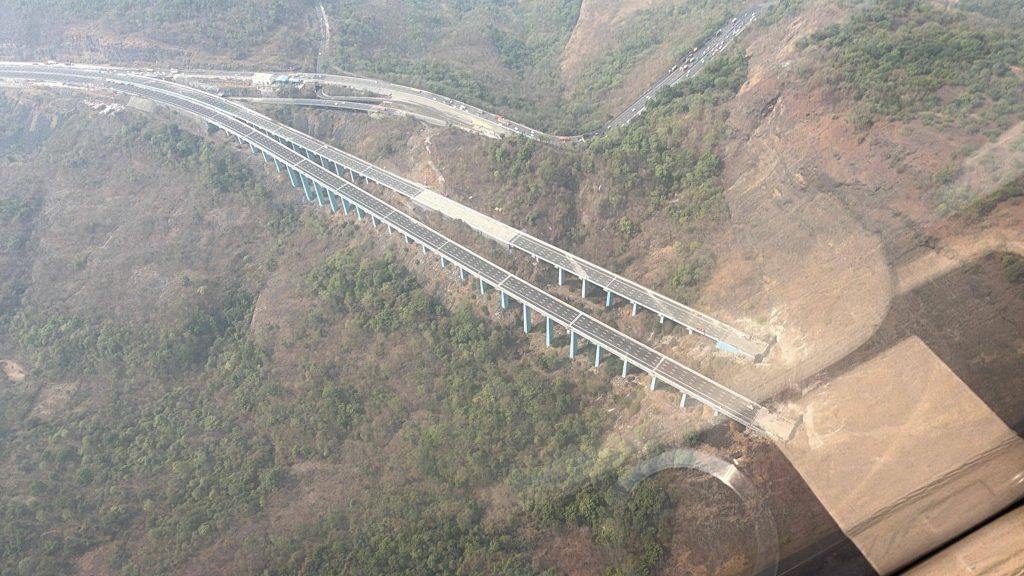 An aerial shot showing the old winding ghat road on the left contrasted with the new, straight multi-lane "Missing Link" bypass and its towering blue-pillared viaduct on the right.