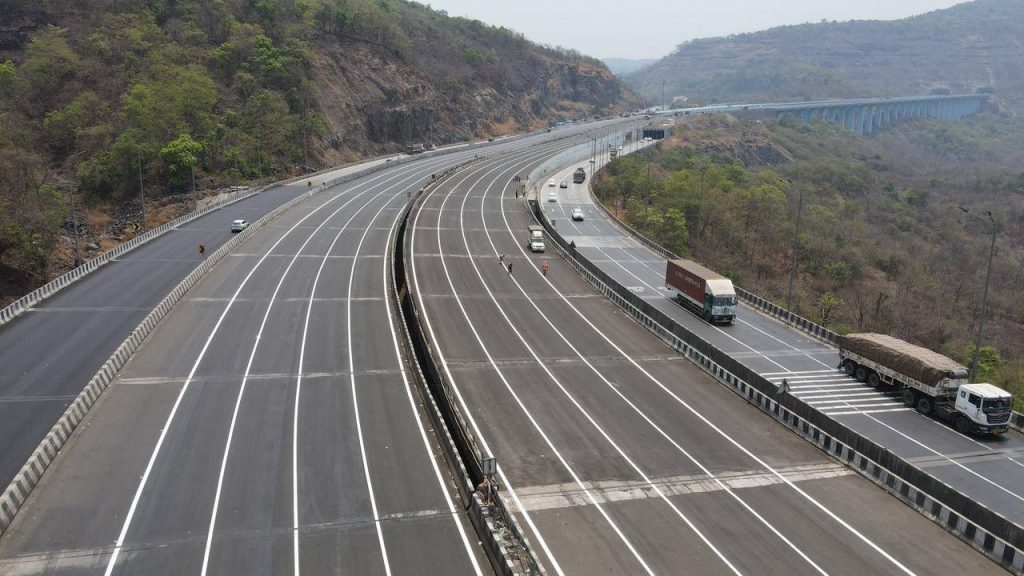 A high-angle view of the expansive 8-lane concrete carriageway of the Mumbai-Pune Expressway "Missing Link," showing a sweeping curve through a mountainous landscape with a long viaduct visible in the background.