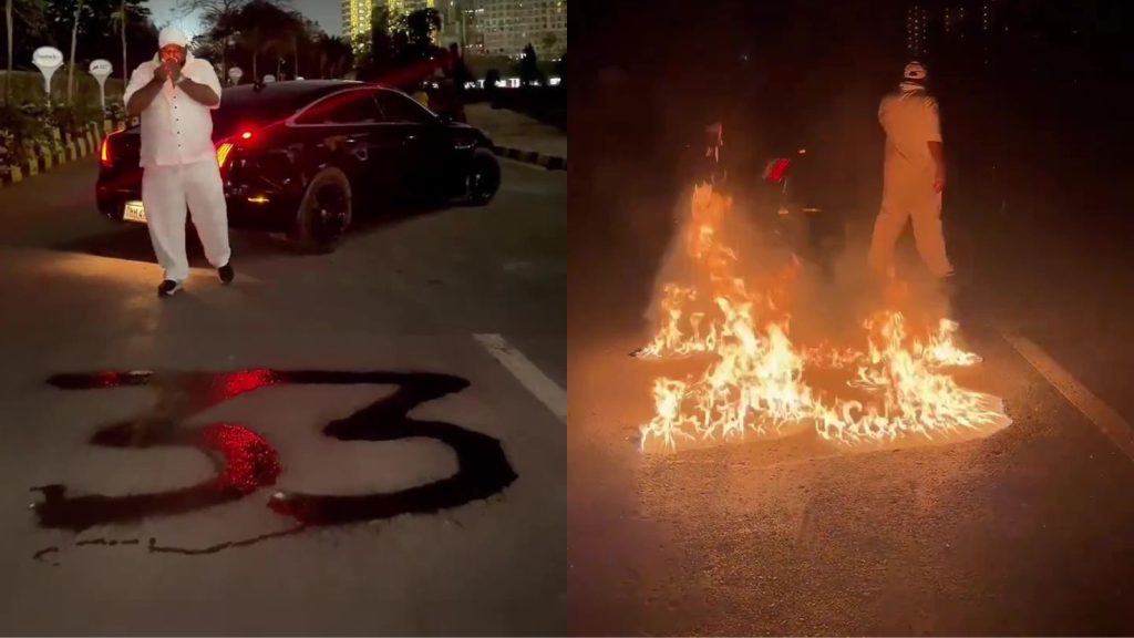 A side-by-side split image showing a man in white clothing performing a dangerous stunt on a dark road at night. On the left, the man stands near a black luxury car while liquid fuel is poured onto the asphalt in the shape of the number "33." On the right, the same number "33" is engulfed in bright orange flames on the road surface as the man walks away in the background. Buildings and streetlights are visible in the distant background under a night sky.