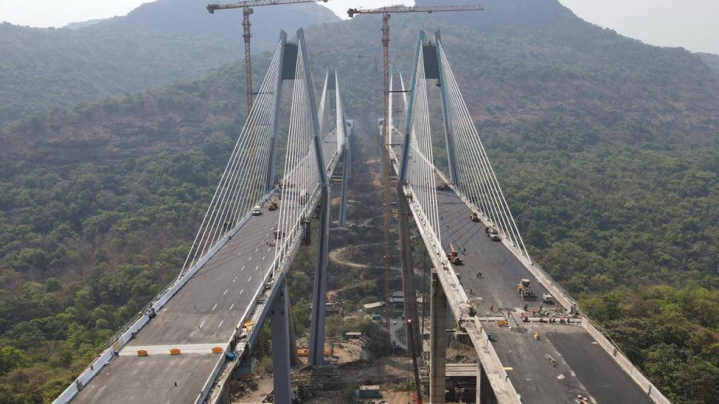 A perspective shot from the bridge deck level, looking down the length of the twin cable-stayed spans with construction cranes and the surrounding mountain peaks in the distance.