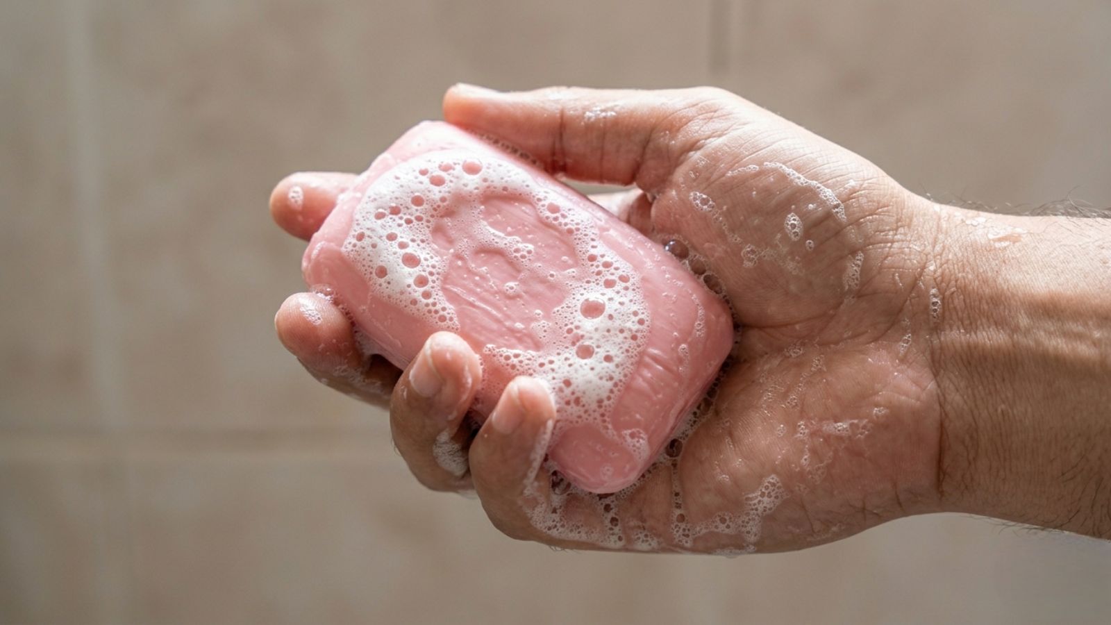 A close-up, wide-angle shot of a person's hand holding a pink bar of bathing soap covered in white foam bubbles, set against a blurred, neutral-toned background.