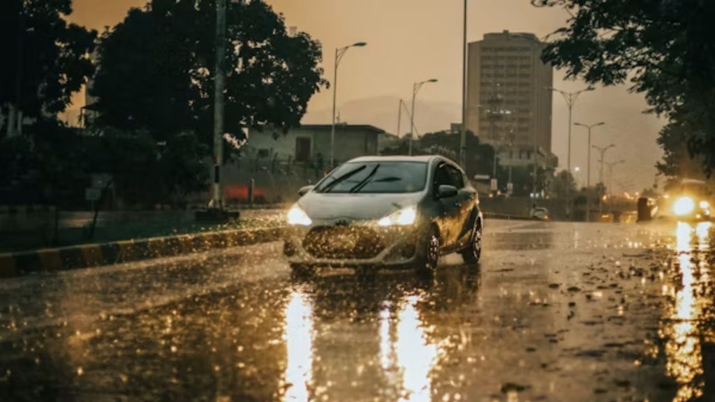 A silver hatchback car driving down a wet city street at dusk during heavy rainfall, with its headlights reflecting strongly on the waterlogged asphalt.