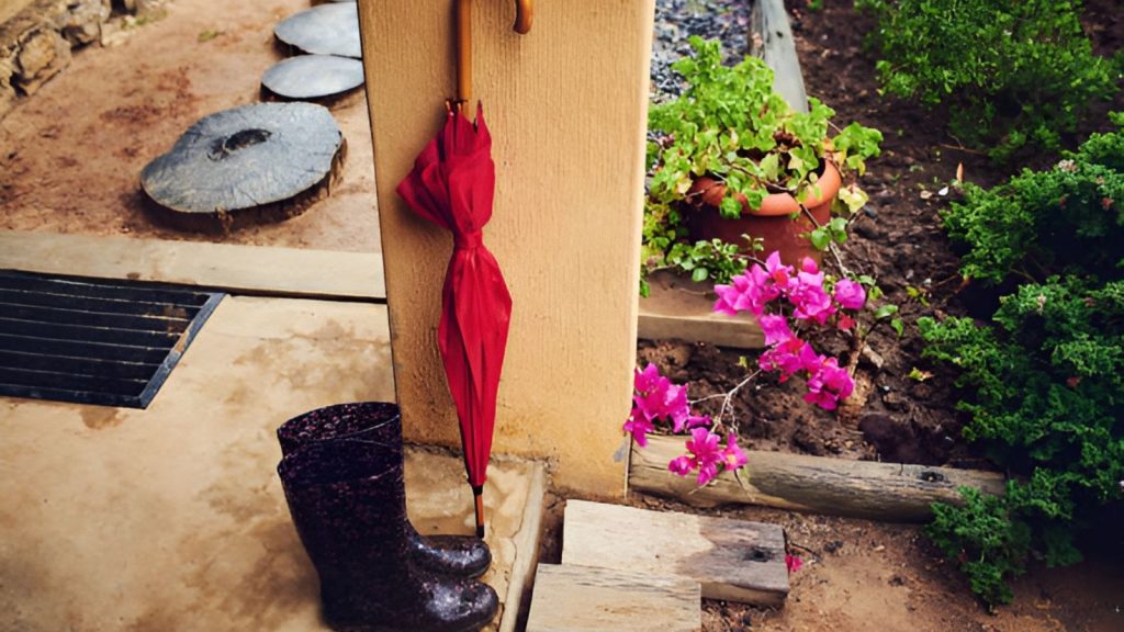A pair of dark, patterned rubber rain boots and a folded red umbrella leaning against a beige porch pillar next to a garden with blooming pink bougainvillea flowers during a rain shower.