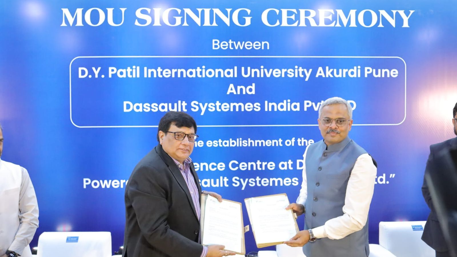 Two officials from D.Y. Patil International University and Dassault Systèmes India stand in front of a blue "MOU SIGNING CEREMONY" backdrop, holding signed folders and shaking hands to commemorate their partnership.