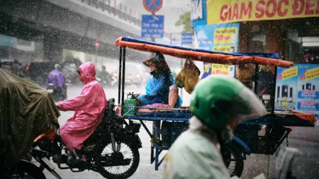 Commuters navigating a busy city street in heavy rain, including a person in a bright pink raincoat on a motorcycle and another individual on a covered motorized cart.