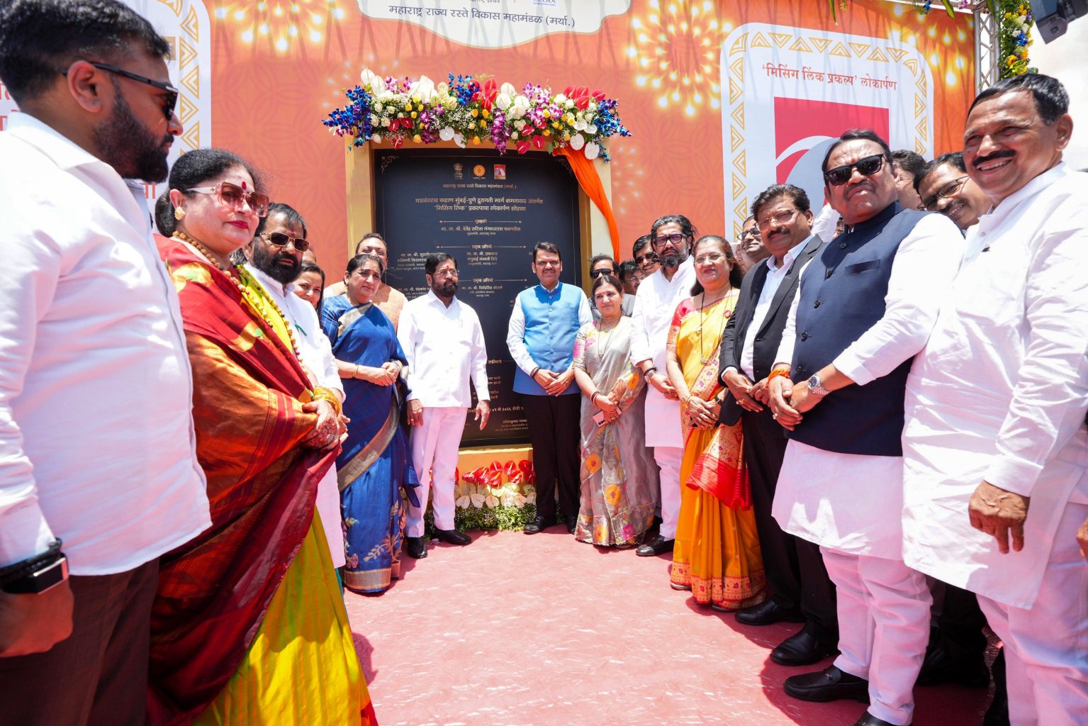 A group of Indian political dignitaries, including Chief Minister Devendra Fadnavis, Deputy Chief Minister Eknath Shinde, and Deputy Chief Minister Sunetra Ajit Pawar, stand smiling together in front of a ceremonial plaque decorated with flowers during the inauguration ceremony of the Missing Link project.
