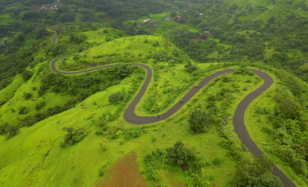 A stunning aerial photograph (drone shot) of a winding, serpentine asphalt road cutting through vibrantly green, rain-washed Sahyadri hills. The complex S-curves of the road are clearly visible, with small houses of a village nestled further up the mountain. Two small vehicles are seen navigating the quiet, twisting path.