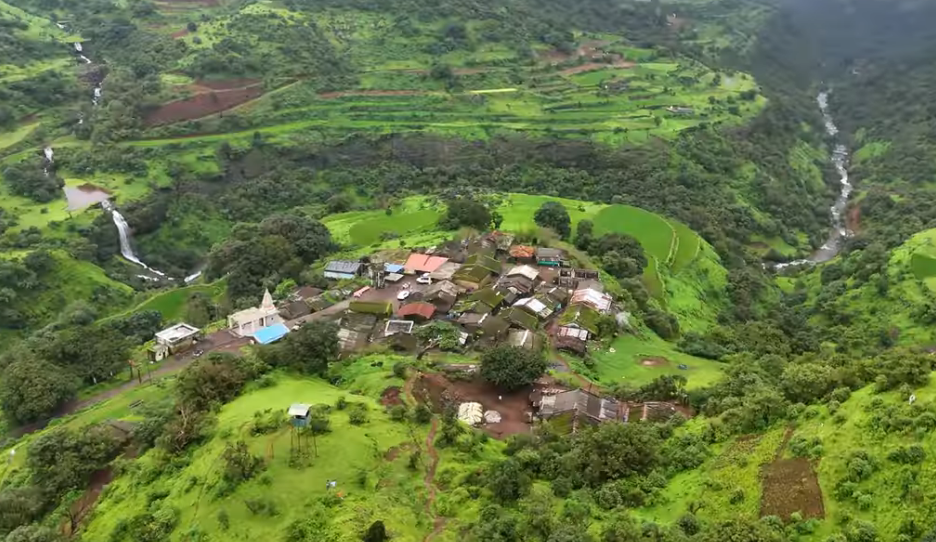 A wide-angle aerial perspective (drone view) showing a compact cluster of simple village houses with tiled roofs nestled on a steep, agricultural terraced hillside. A small, white-spired temple sits slightly apart. Numerous waterfalls cascade down the deep gorge opposite the village. The surrounding landscape is an expansive carpet of lush, vibrant green hills under a cloudy sky.