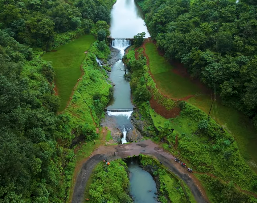 A serene landscape photograph of a multi-tiered waterfall cascading down a rocky gorge. The surrounding hills are covered in vibrant, lush green vegetation characteristic of Phopsandi during the monsoon. A simple bridge is visible in the upper background across the stream. The foreground features textured, wet rock formations, framing the natural water feature.