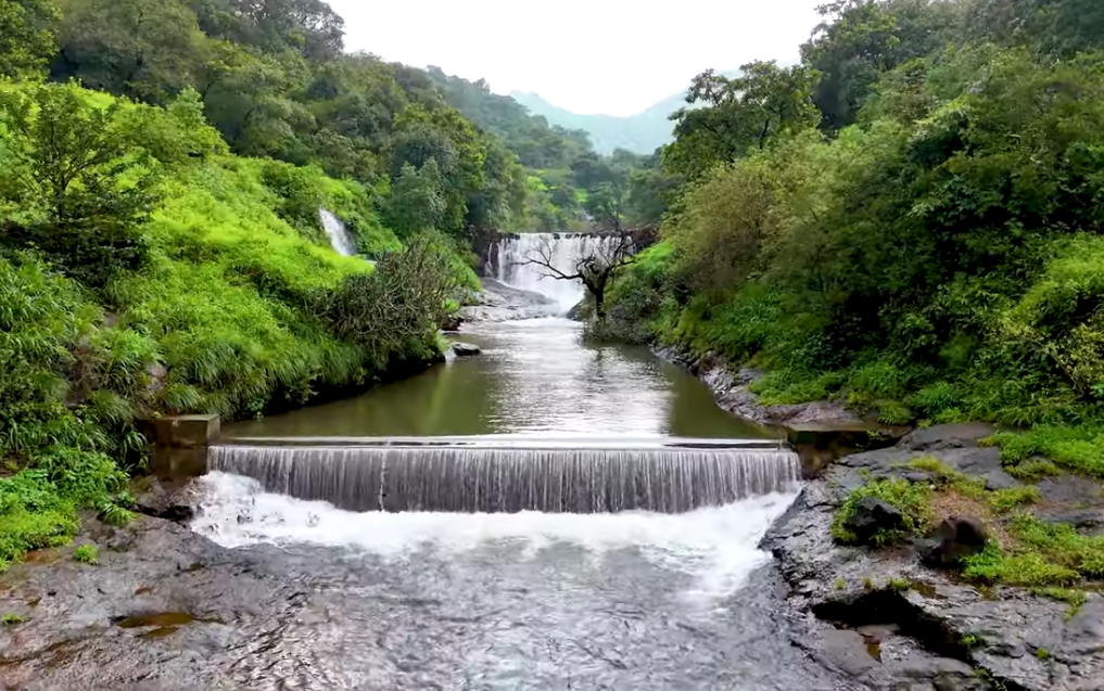 A peaceful landscape view of a slow-moving river or reservoir with a small, concrete check dam creating a gentle overflow in the foreground. The river is surrounded by dense, lush green forests and vegetation, leading up to mist-covered hills in the distance. The overcast conditions create a serene and mystical atmosphere.