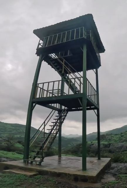 A vertical photograph looking up at a tall, green-painted metal watchtower (machan). The tower is built on a concrete base with two platforms and a gabled roof, accessed by steel stairs. The background shows a vast, rain-soaked valley with rolling green hills and a cloudy, overcast sky, emphasizing the tower's function as a viewpoint for panoramic Sahyadri vistas.