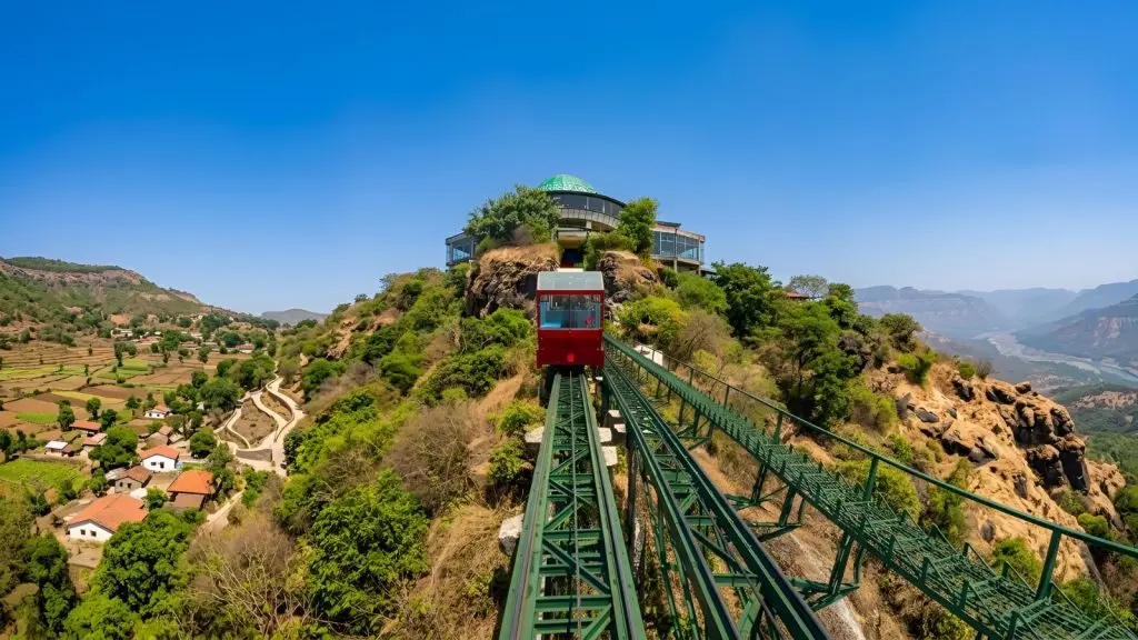 A wide 16:9 panoramic view of a steep funicular railway track ascending a lush mountain ridge near Mahabaleshwar. A bright red railway car travels up the green steel tracks toward a prominent, dome-roofed revolving restaurant perched at the summit. The landscape opens up on either side, showing a terraced village with red-roofed houses nestled in the valley to the left and sprawling, sunlit mountain ranges carving through the terrain to the right under a clear blue sky. The place is called - Vidnyangad Tarangan.