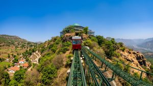 A wide 16:9 panoramic view of a steep funicular railway track ascending a lush mountain ridge near Mahabaleshwar. A bright red railway car travels up the green steel tracks toward a prominent, dome-roofed revolving restaurant perched at the summit. The landscape opens up on either side, showing a terraced village with red-roofed houses nestled in the valley to the left and sprawling, sunlit mountain ranges carving through the terrain to the right under a clear blue sky. The place is called - Vidnyangad Tarangan.