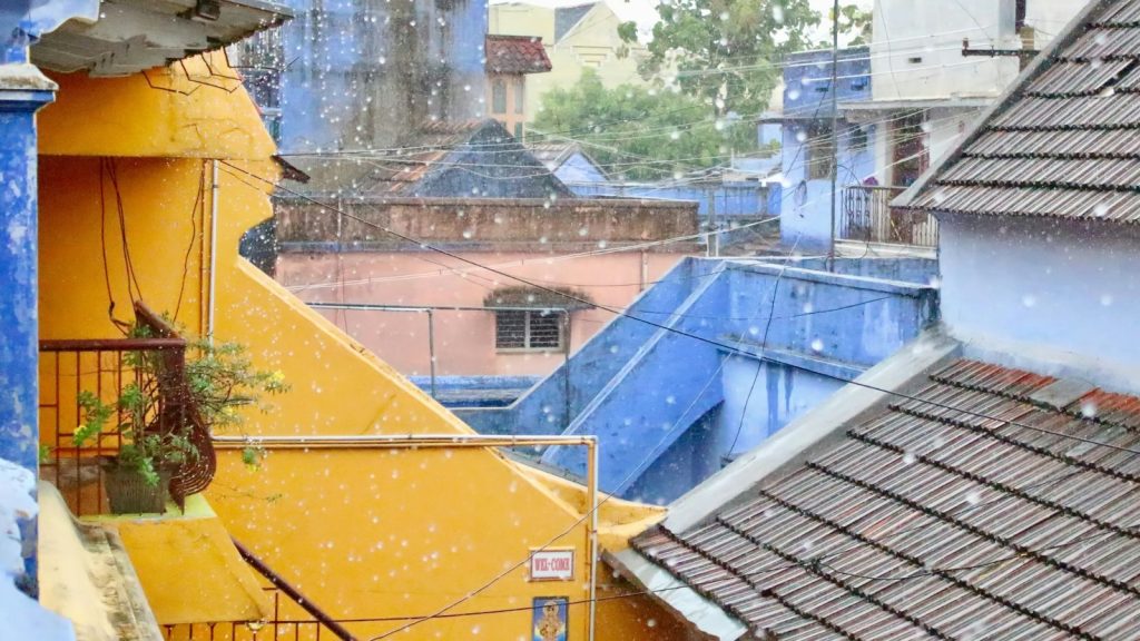 A high-angle view of closely packed urban houses with yellow and blue painted walls and tiled roofs during a heavy monsoon downpour.