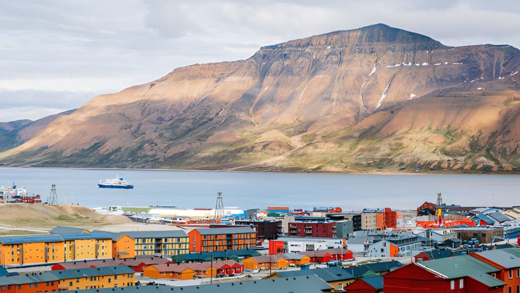 An elevated, panoramic view of a colorful Arctic town with rows of orange, yellow, red, and blue apartment blocks and buildings along a waterfront. A large ship sails in the calm bay, and massive, barren, snow-dappled mountains rise steeply in the background under an overcast sky.