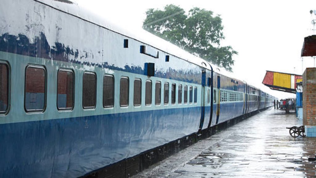 A long blue and light-blue Indian passenger train parked at a wet, glistening railway station platform during a daytime downpour.