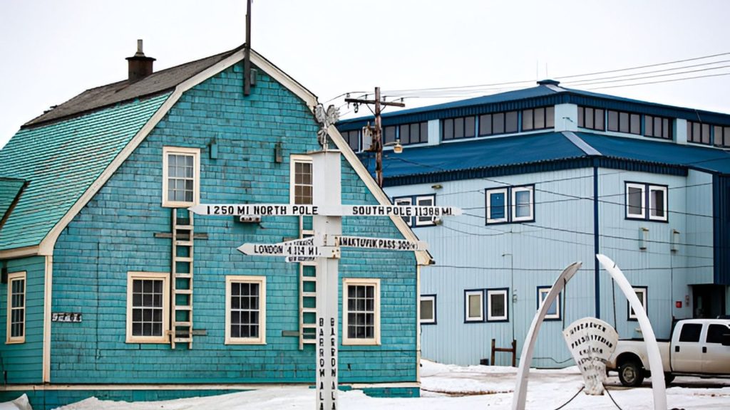 A street view in Utqiagvik, Alaska, featuring a white wooden multi-directional signpost in the foreground. The signs point toward geographic locations, including "1250 MI NORTH POLE" and "SOUTH POLE 11388 MI." In the background stand a teal shingled wooden building with an attached exterior ladder and a large light-blue industrial structure under a bright, overcast sky.