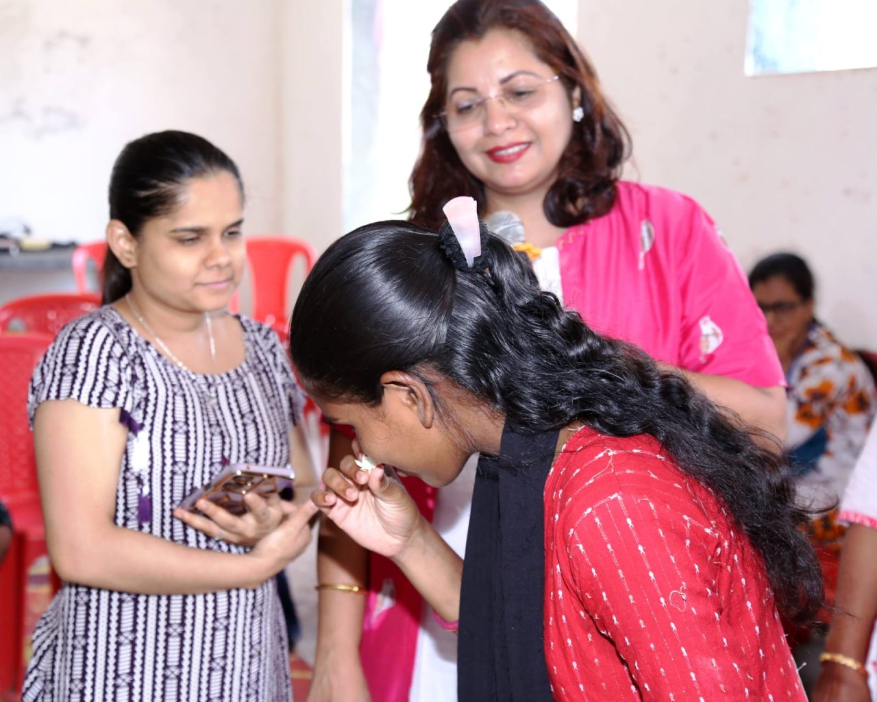 A visually impaired student with long dark hair, wearing a red top and black dupatta, smiles as she brings a white jasmine flower to her nose. Behind her, Dr. Meghana Jalgaonkar in a bright pink jacket smiles, and another woman in a patterned dress holds a smartphone during the sensory photography workshop in Virar.