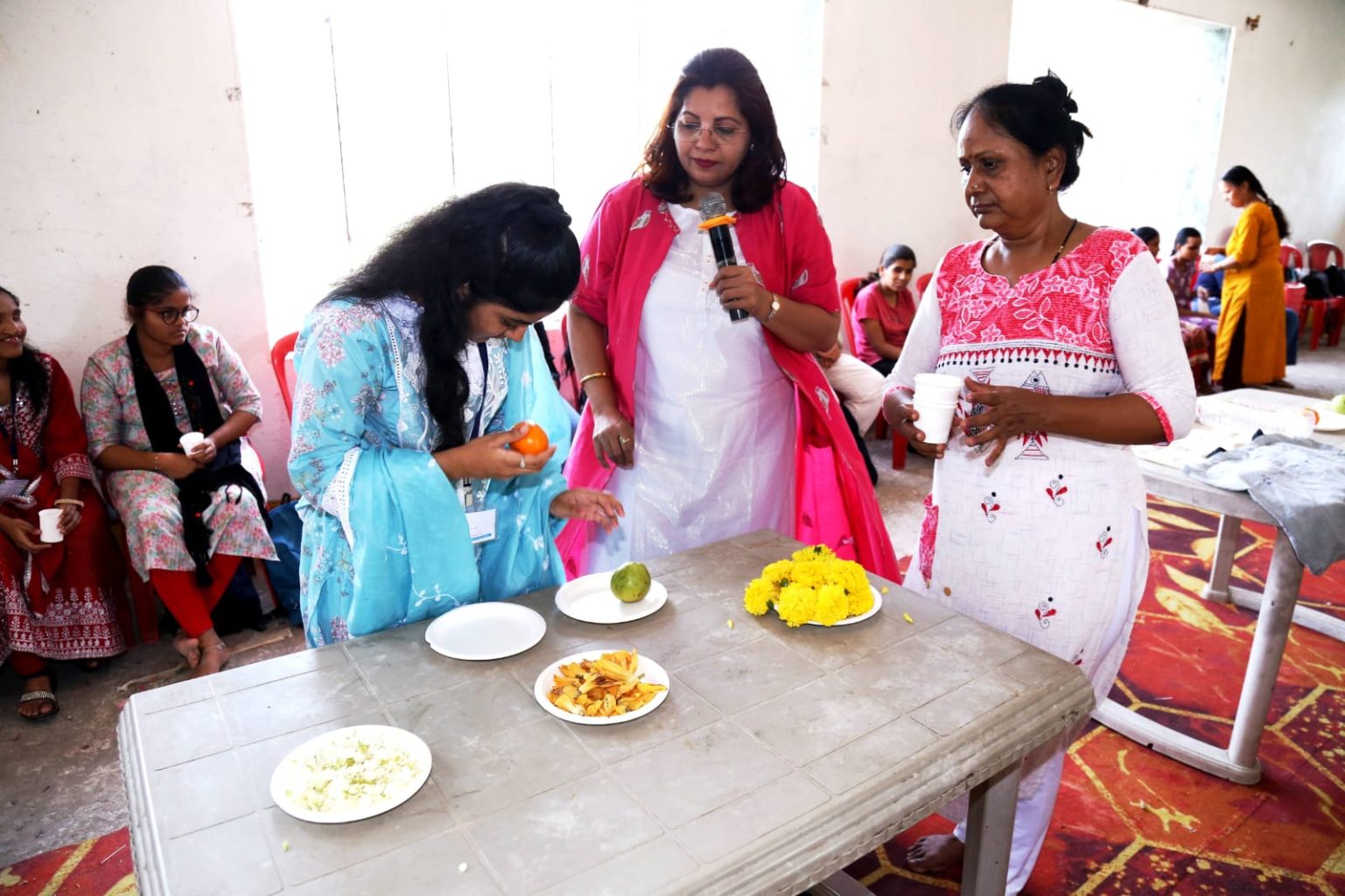 A visually impaired student in a blue outfit carefully examines an orange with her hands to understand its texture and shape. Dr. Meghana Jalgaonkar and an assistant look on as the student explores a table filled with sensory items like fruits, flowers, and snacks during the Sparsh Shakti residential camp.