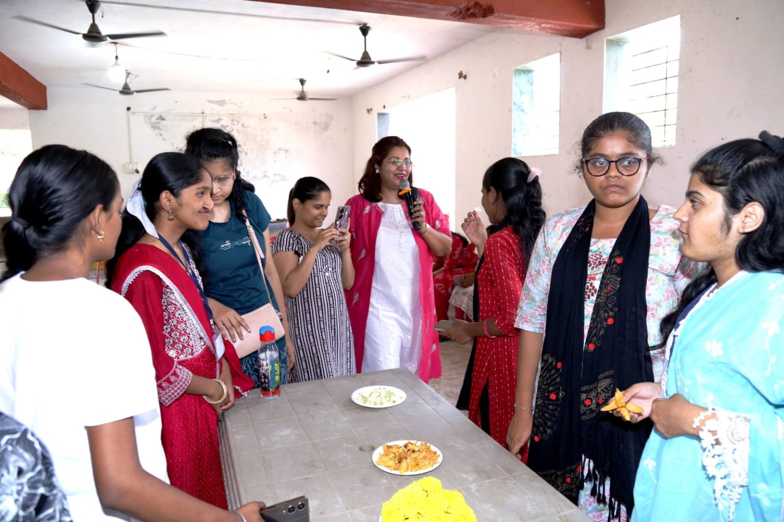 A group of visually impaired female students gathered around a table featuring plates of fragrant flowers during the Sparsh Shakti workshop. Dr. Meghana Jalgaonkar, holding a microphone, facilitates the session while students interact with the materials to learn about distance and perception through smell.