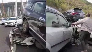 A side-by-side split image from the file Accident on the Newly Opened Missing Link.jpg showing the aftermath of a severe multi-vehicle pile-up on a highway bridge. On the left, a dark-colored SUV is seen severely crushed on top of a white sedan, with its rear tires completely elevated off the ground by the crumpled hood of the car beneath it. On the right, the side view of a silver SUV shows it wedged tightly against the wreckage, with a man standing nearby surveying the severe engine bay damage amidst heavy highway traffic and a cable-stayed bridge structure visible in the background.