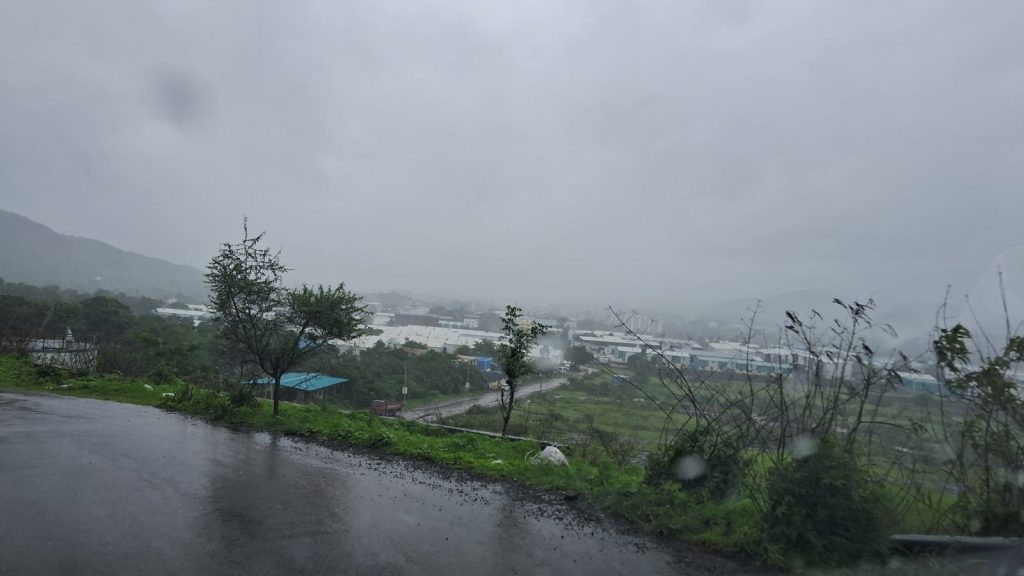 A wide landscape view on a rainy, overcast day in Pune. In the foreground, a wet, dark asphalt road curves along a green hillside. Below, a misty valley reveals industrial or residential buildings with white rooftops surrounded by lush green trees and fields. The entire scene is shrouded under a heavy, grey, monsoon sky with visible raindrops blurring the frame, capturing a fresh and gloomy monsoon atmosphere.