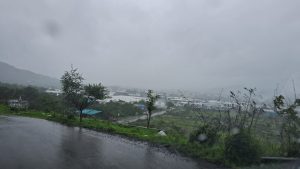 A wide landscape view on a rainy, overcast day in Pune. In the foreground, a wet, dark asphalt road curves along a green hillside. Below, a misty valley reveals industrial or residential buildings with white rooftops surrounded by lush green trees and fields. The entire scene is shrouded under a heavy, grey, monsoon sky with visible raindrops blurring the frame, capturing a fresh and gloomy monsoon atmosphere.