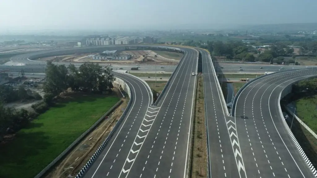 Aerial view of a modern, multi-lane access-controlled expressway interchange featuring sweeping curved flyovers, crisp white road markings, and clear directional lane arrows, representing India's high-speed highway infrastructure under a clear sky.