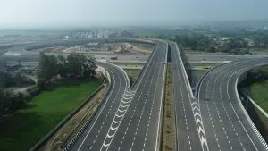 Aerial view of a modern, multi-lane access-controlled expressway interchange featuring sweeping curved flyovers, crisp white road markings, and clear directional lane arrows, representing India's high-speed highway infrastructure under a clear sky.