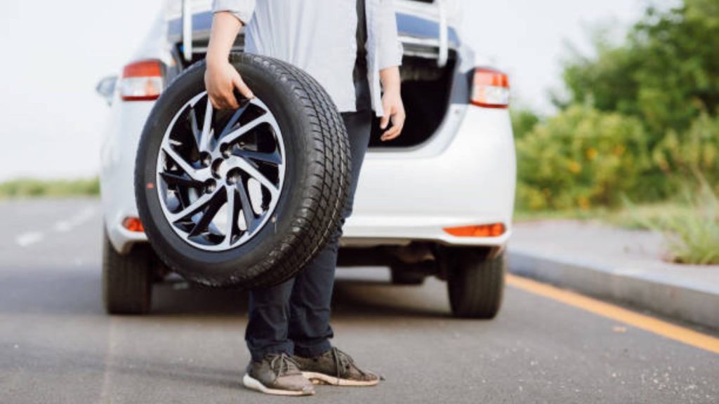 A person standing on the side of a highway next to a white car with its trunk open, holding a spare tire with a black and silver alloy wheel rim, illustrating a vehicle breakdown or puncture repair situation.